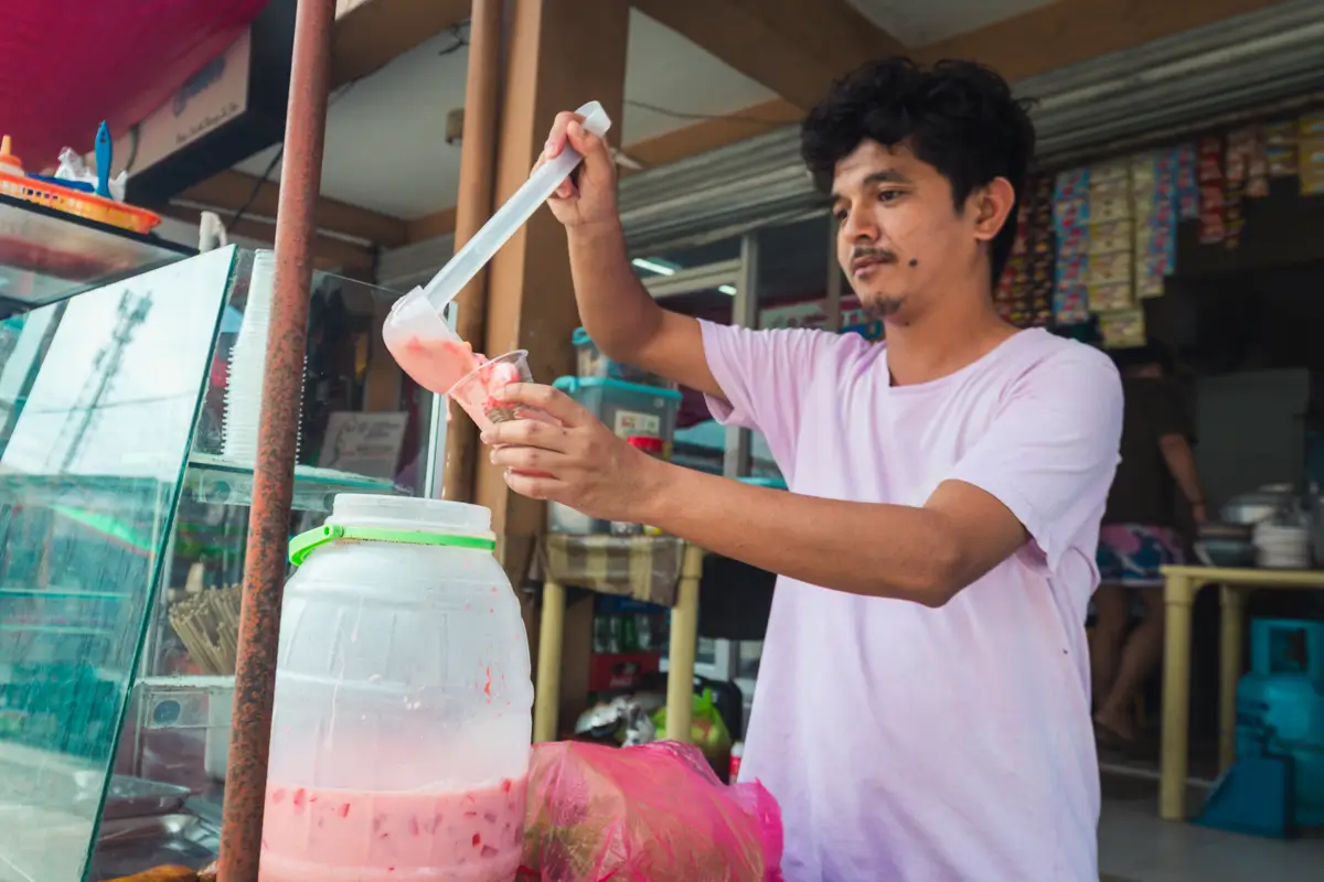 A man in a light pink shirt scoops a pink drink from a large plastic container into a cup at an outdoor food stall in Moalboal. Shelves with packaged goods are visible in the background.