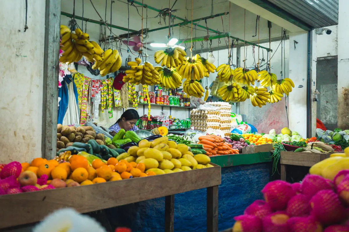 A bustling market stall in Moalboal displays bananas, mangoes, oranges, apples, and assorted vegetables. Packaged goods line shelves behind the stall, while a person stands behind the counter, partially hidden by fresh produce.