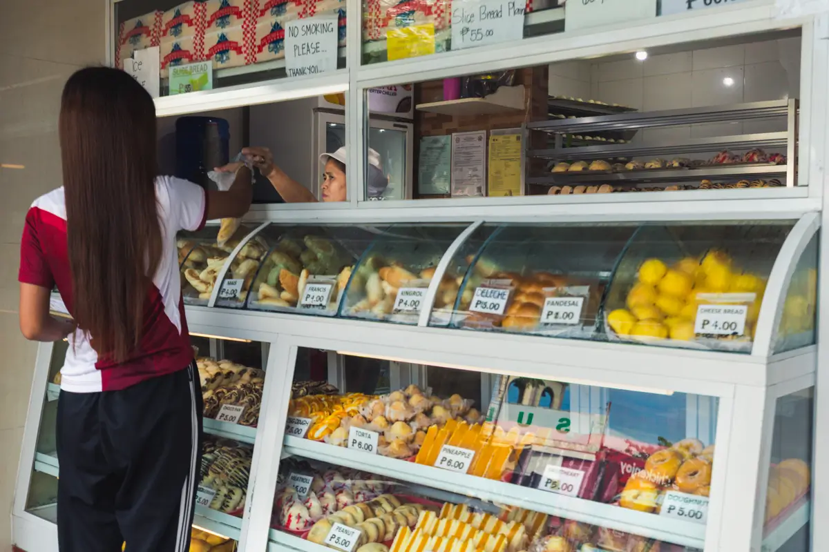A woman stands in front of a Moalboal bakery display case filled with assorted breads, pastries, and packaged snacks, while handing money to a cashier behind the counter. Prices are visible on the glass.