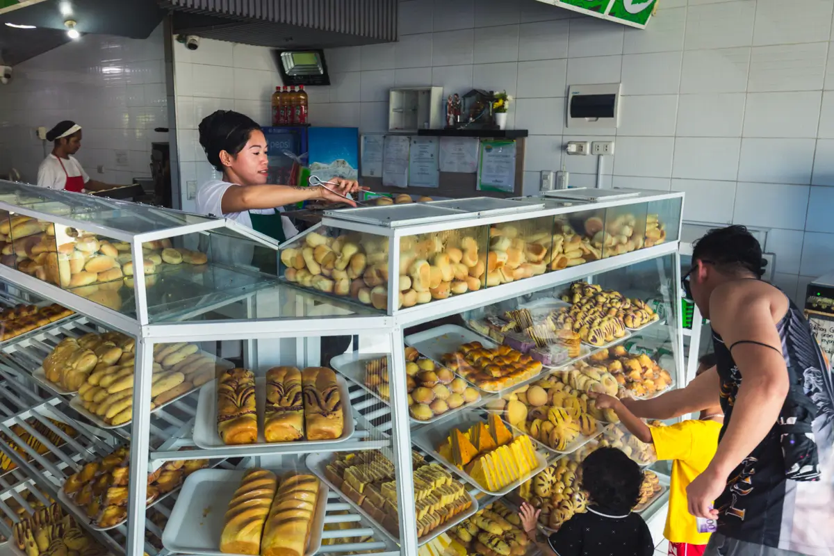 A bakery in Moalboal with glass display cases filled with various breads and pastries; a staff member serves customers, including an adult and two children, while another worker is in the background.