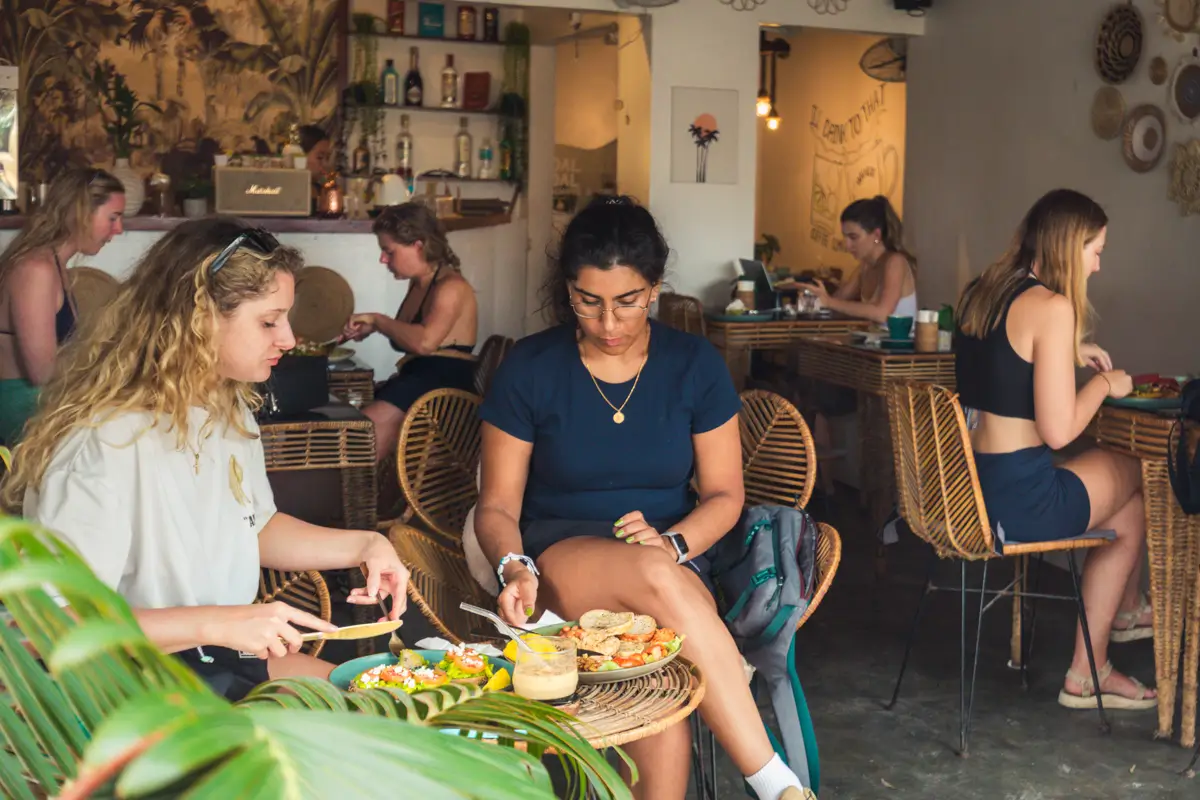 Several people sit at wicker tables in a cozy Moalboal café, eating and talking. Two women in the foreground focus on their meals, while shelves with bottles and a small bar area are visible in the background.