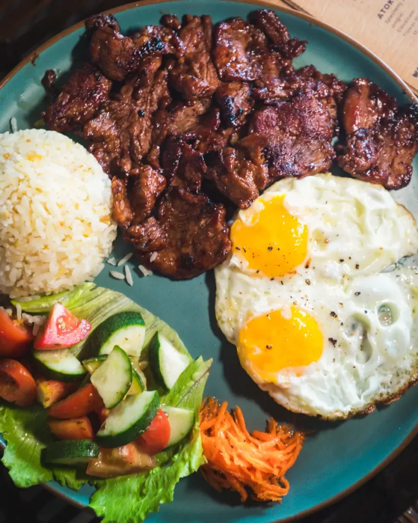 A plate inspired by Moalboal, featuring grilled marinated meat, two sunny-side-up eggs, a mound of white rice, and a fresh side salad with lettuce, cucumber, tomato, and shredded carrot.
