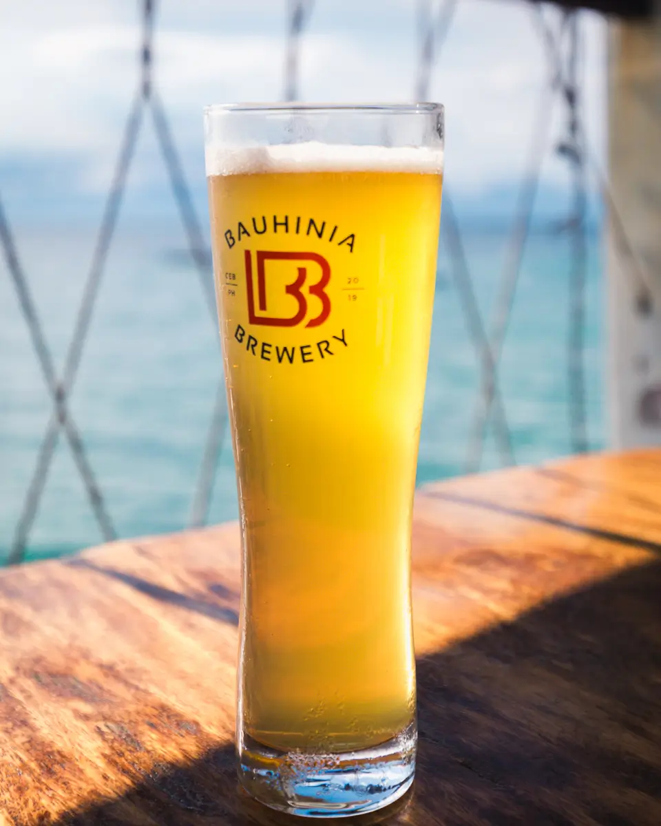 A tall, clear glass of light yellow beer with a foamy head, labeled Bauhinia Brewery, sits on a wooden table in Moalboal. The background shows an out-of-focus view of water and part of a railing.