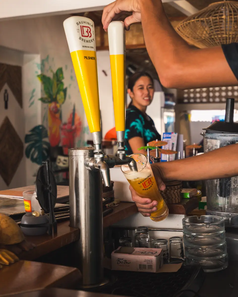A person fills a tall glass with beer from a tap at a Moalboal bar counter while a woman stands and smiles in the background. The bar area features stacked glasses, a blender, and colorful wall art.