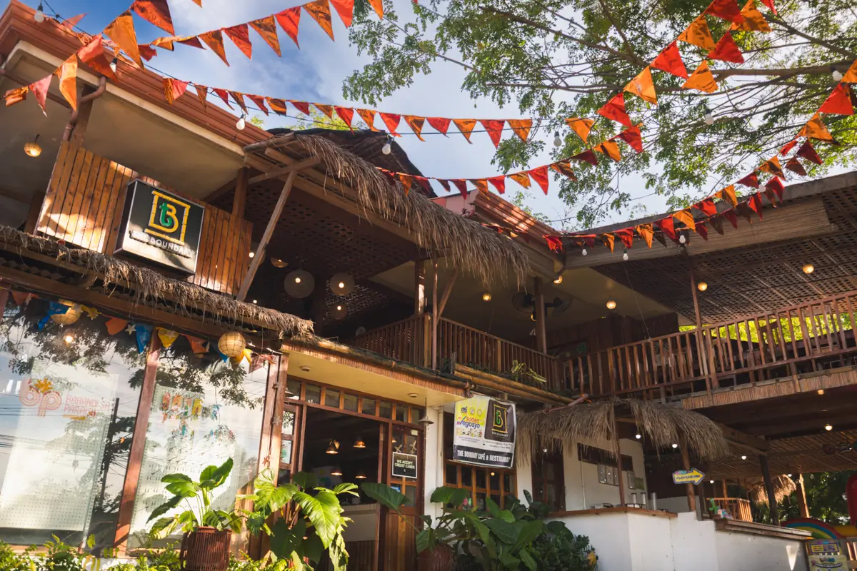 A two-story restaurant in Moalboal with wooden and bamboo elements, thatched roof accents, and string lights. Orange and red pennant banners hang above, with green plants in front of the building. The sky is partly cloudy.