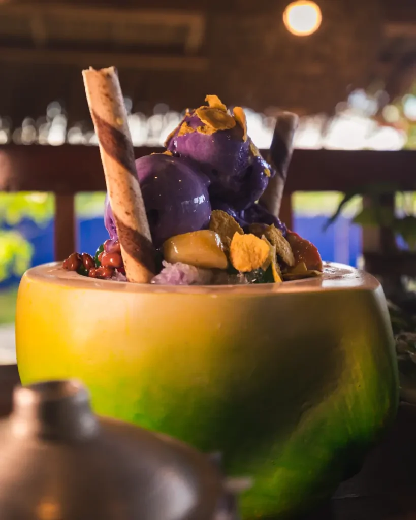 A close-up of a halo-halo dessert served in a green coconut shell in Moalboal, topped with purple ice cream, wafer sticks, fruit pieces, and cereal flakes, with a blurred background and warm lighting.