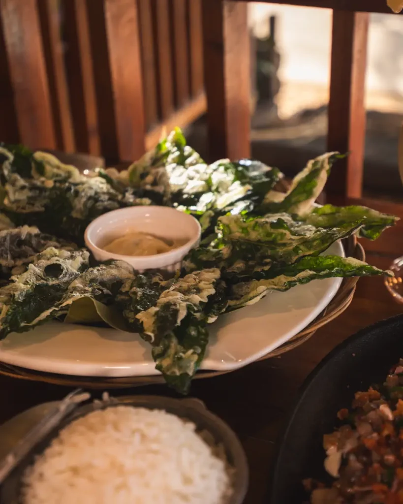 A plate of crispy fried green leaves, a nod to Moalboal flavors, encircles a small cup of dipping sauce, with white rice and a portion of chopped meat dish partially visible in the foreground.