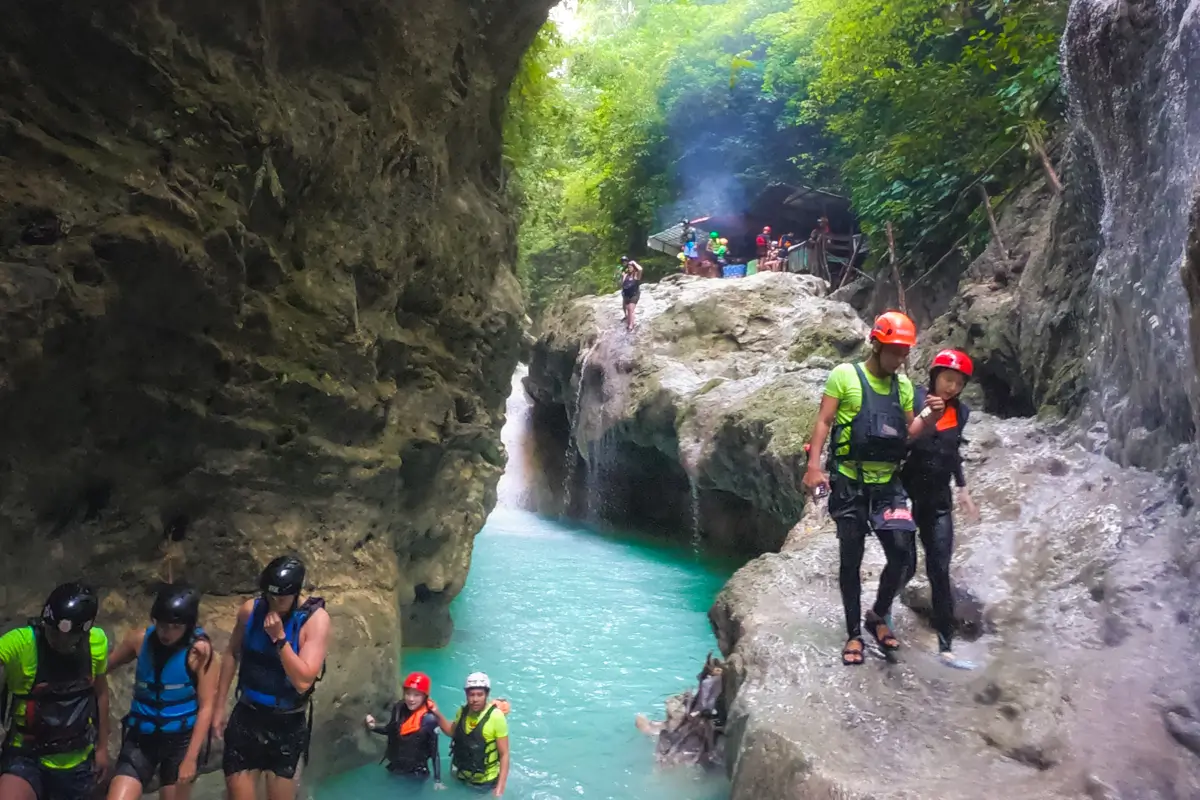 People in helmets and life vests are canyoning in a rocky gorge with turquoise water near Moalboal. Some stand on rocks, others wade through the stream as dense green foliage surrounds them and smoke rises in the background.