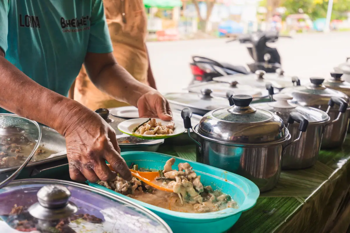 A person serves food from a tray into a plate at a Moalboal food stall, with several covered pots and containers arranged on a table outdoors.