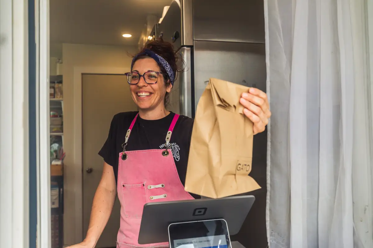 A woman wearing glasses and a pink apron stands behind a counter in Fernie, smiling as she holds out a brown paper bag. A point-of-sale device sits in front of her, with a white curtain to the right.