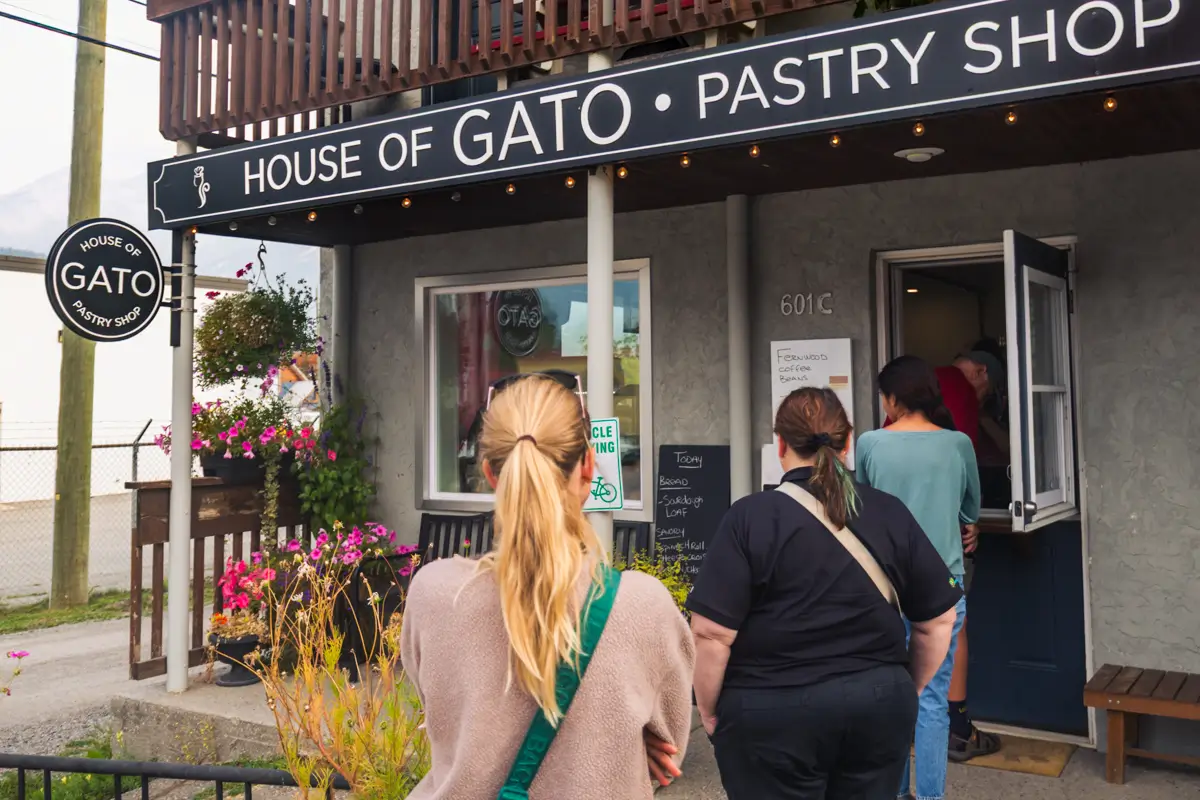 Several people stand in line outside the entrance of House of Gato Pastry Shop in Fernie, a gray building with a black sign. Potted flowers decorate the storefront, and a menu is posted beside the open door.