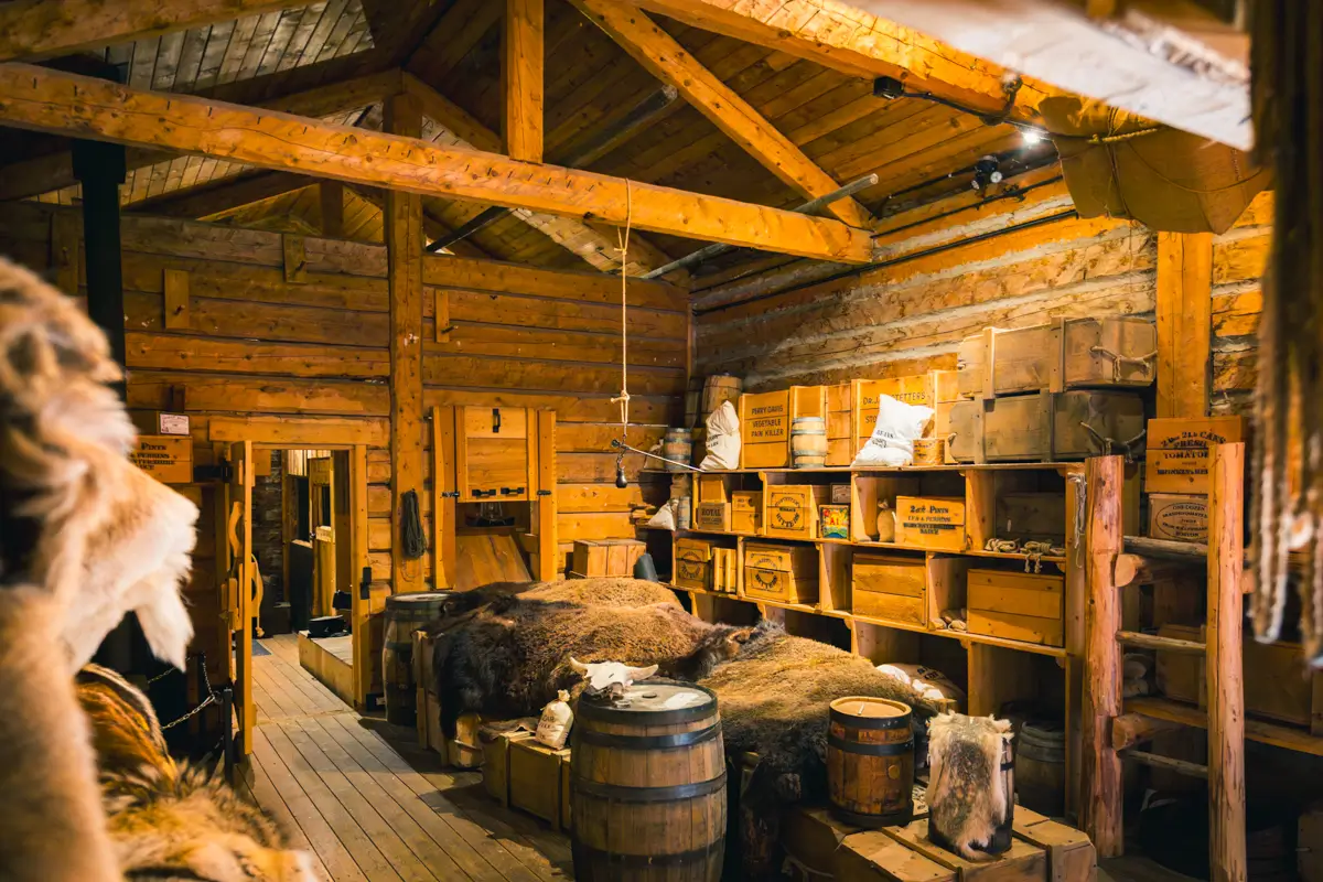 Interior of a log cabin in Lethbridge with wooden beams, stacked crates, animal pelts on a bed, barrels, and various goods on shelves. Warm lighting creates a rustic, historical atmosphere.