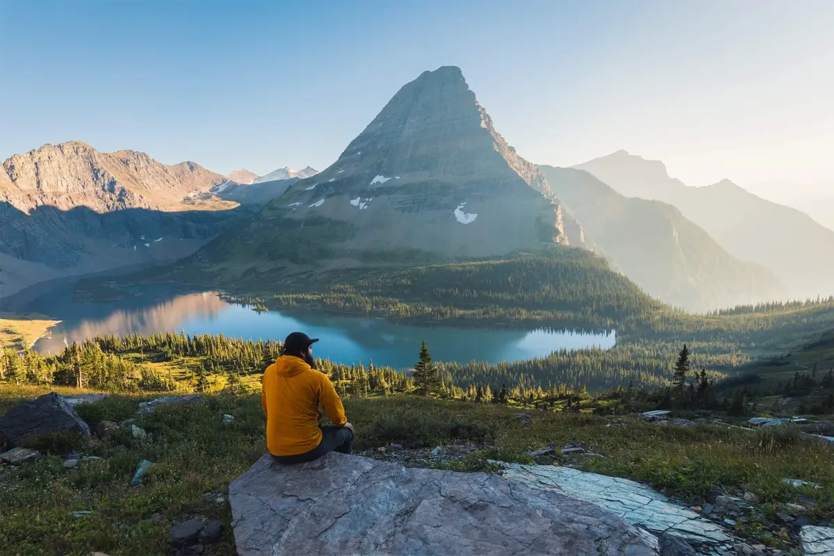A person in a yellow jacket sits on a rock, overlooking a lake and pine trees with a large mountain in the background under a clear sky, evoking the natural beauty found near Lethbridge.