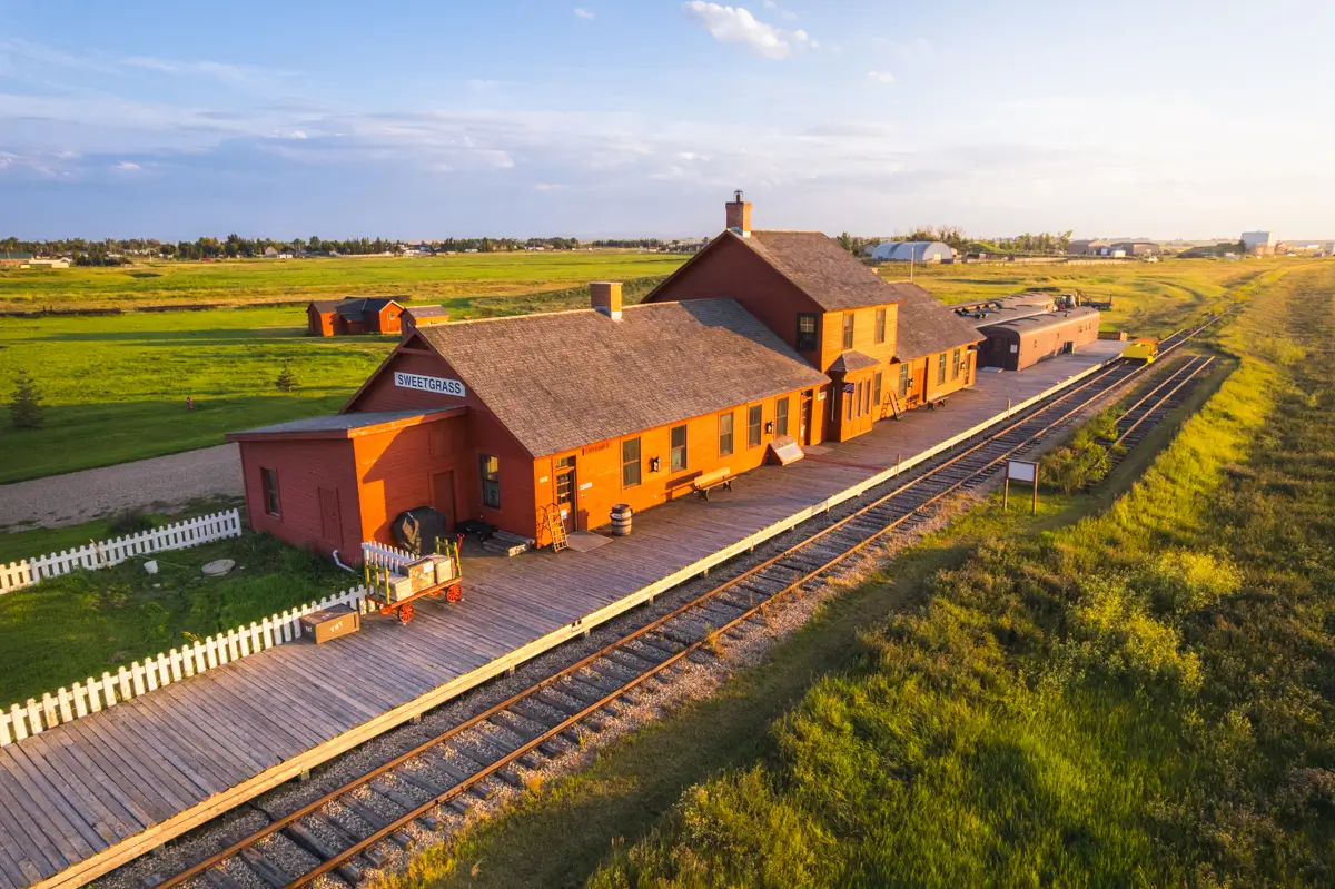 A historic Lethbridge train station with a wooden platform and railway tracks runs through a grassy landscape under a clear sky. The red building is accented by a white picket fence lining part of the property.