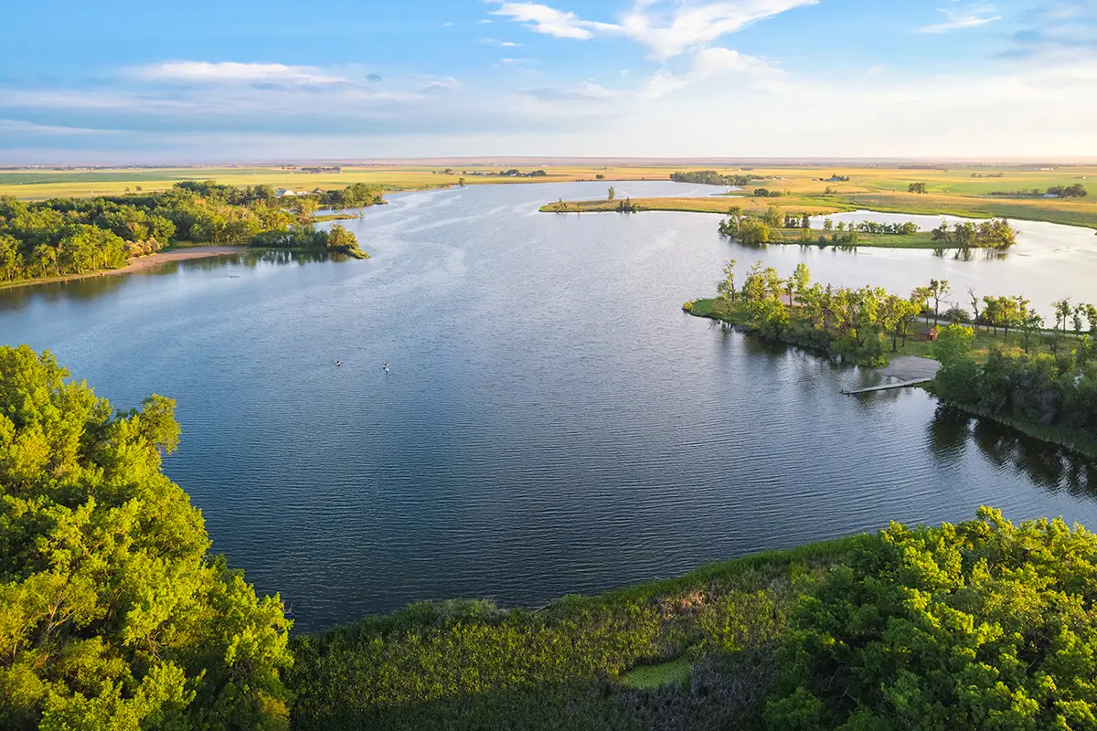 A wide river or lake near Lethbridge with small tree-covered islands, a grassy shoreline, and open fields in the background beneath a blue sky with scattered clouds. The calm water is surrounded by lush green vegetation.