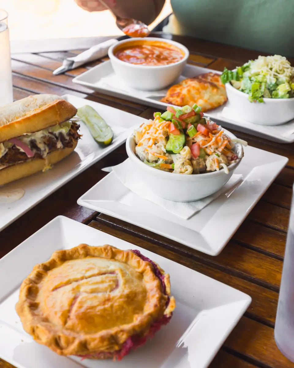 A wooden table in Lethbridge is set with plates of food, including a sandwich with a pickle, a bowl of pasta salad with diced vegetables, a bowl of soup, a small pie, and a salad. A person is seated in the background.