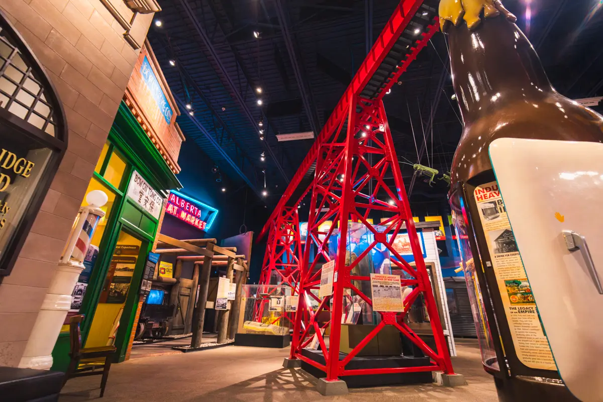 Indoor Lethbridge exhibit featuring a large red metal structure, colorful storefronts on the left, and a giant bottle display on the right, all beneath a high, dark ceiling with exposed lights and beams.