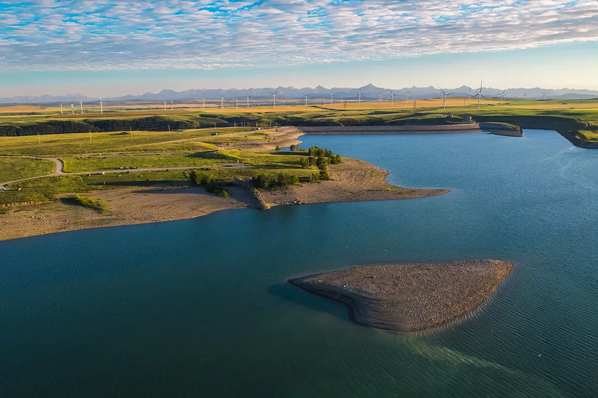 A large reservoir with a rocky shoreline and a small island near Lethbridge, surrounded by grassy fields and distant wind turbines under a partly cloudy sky.