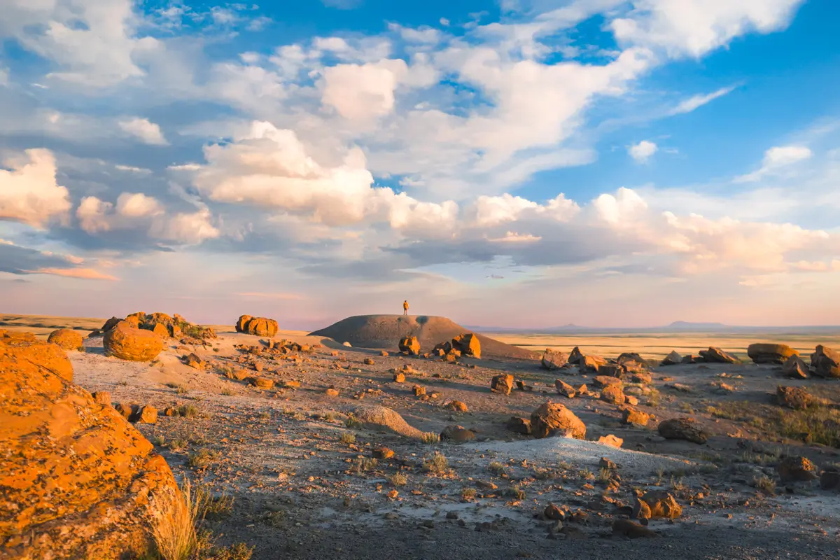 A person stands alone on a small hill in a rocky desert landscape near Lethbridge, under a sky with scattered clouds and bright sunlight. Large orange rocks are scattered across the dry ground.