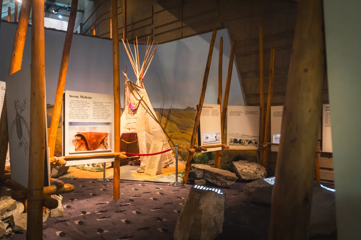 A museum exhibit in Lethbridge displays a model of a tipi surrounded by wooden poles, rocks, and informational panels, set against a backdrop of grassy landscape beneath a blue sky.