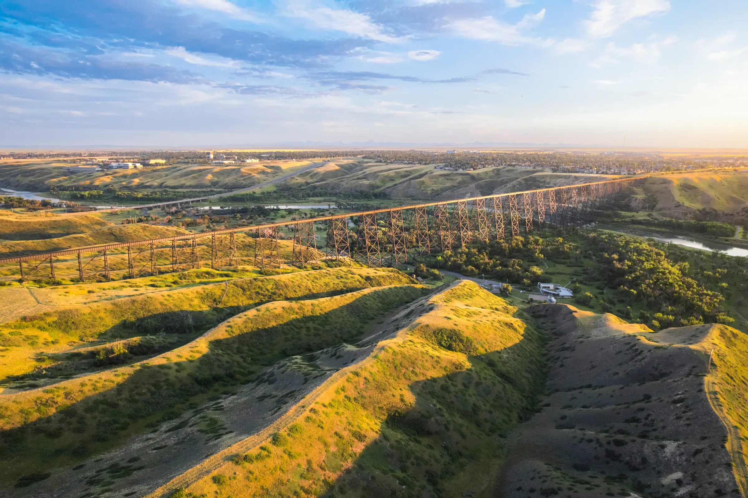 Aerial view of the long steel railway bridge in Lethbridge, stretching across rolling green hills and a winding river, with scattered trees and a partly cloudy sky at sunset.