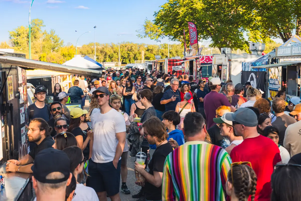 A large crowd gathers outdoors at a Saskatoon food truck festival on a sunny day, standing in lines, socializing, and ordering food from various trucks—one of the top things to do under green trees and a clear blue sky.