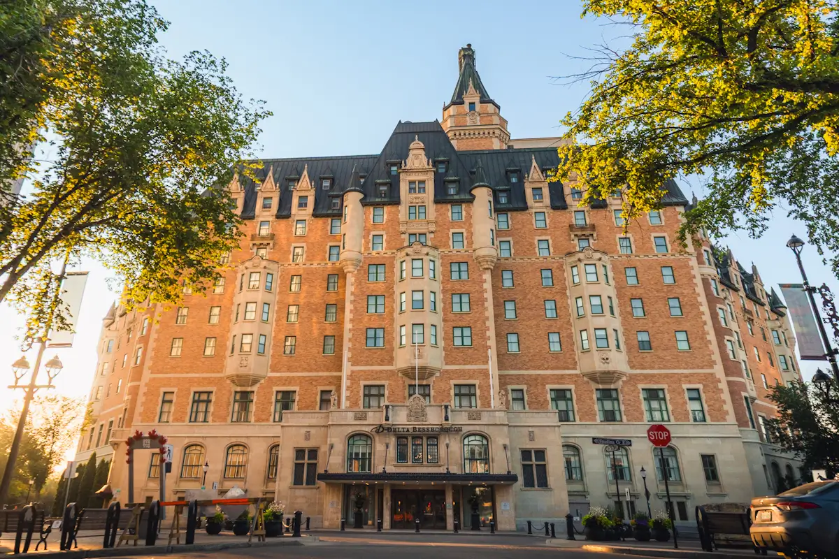 A large, historic hotel with a steep roof and central tower stands under a clear sky, surrounded by trees and sunlight. The entrance reads Delta Hotels Bessborough—an iconic highlight among Saskatoon attractions featured in any Saskatoon guide.