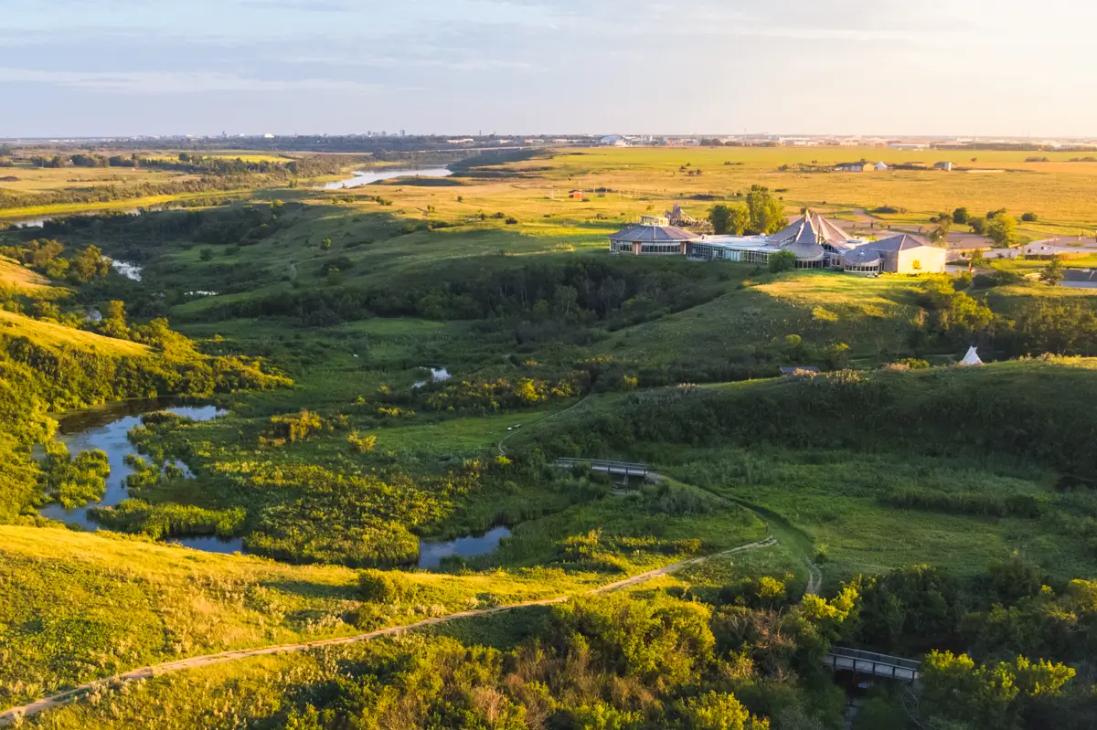 Aerial view of a green landscape with winding trails, small ponds, and scattered trees. A large building complex sits on a hill, open fields stretch out, and the distant city skyline hints at Saskatoon attractions waiting to be explored.