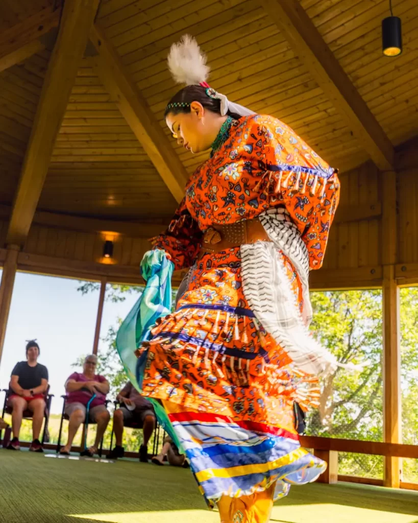 A person in bright orange, beaded traditional Native American regalia dances inside a wooden pavilion while several people watch. Sunlight filters through the windows and trees are visible outside—a highlight among Saskatoon Activities.