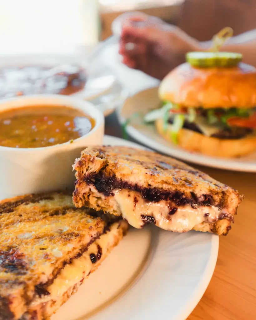 A grilled sandwich with melted cheese and jam is on a plate, beside a cup of soup and a blurred hamburger in the background—a tasty pause during your Saskatoon attractions adventure.