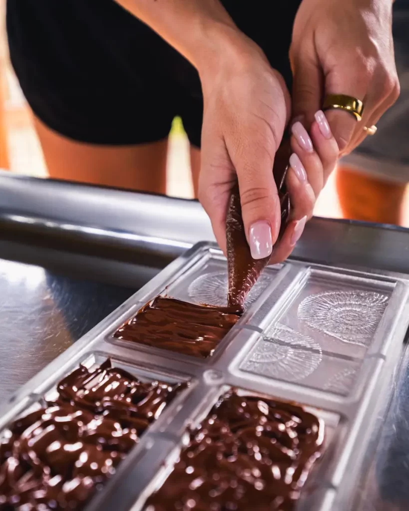 A person squeezes melted chocolate from a piping bag into a rectangular mold; their hands, adorned with neatly manicured nails and a gold ring, craft treats inspired by Bohol’s rich chocolate-making tradition.