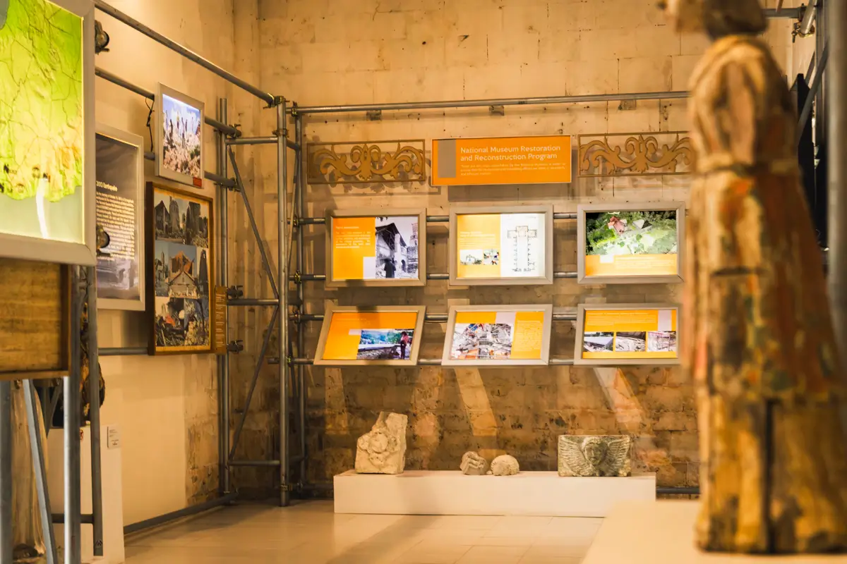 Exhibit displays with images, text panels, and artifacts are arranged on a museum wall made of light-colored Bohol stone. A carved statue stands in the foreground to the right, adding depth to the scene.