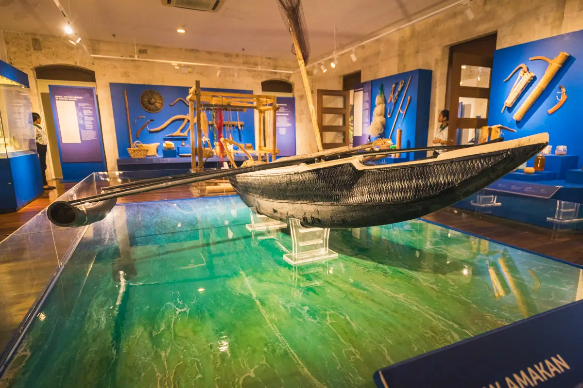 A traditional wooden Bohol boat is displayed on clear stands in a museum exhibit room with blue walls, educational placards, and other cultural artifacts in the background.