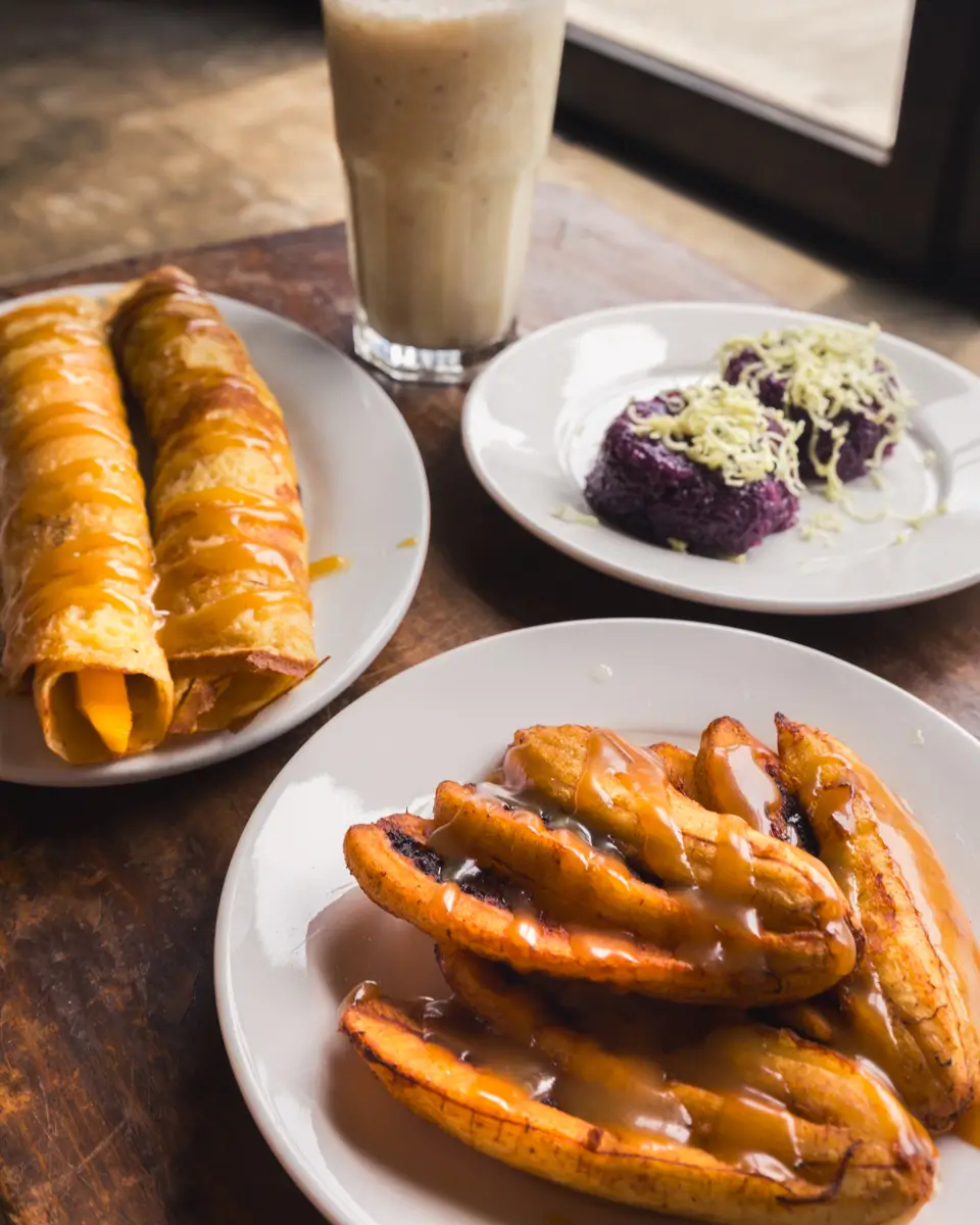 Three white plates with Filipino desserts: two mango-filled rolled crepes with sauce, sliced fried plantains with syrup, and two servings of purple rice cake with grated cheese—served alongside a tall iced drink, evoking sweet flavors from Bohol.