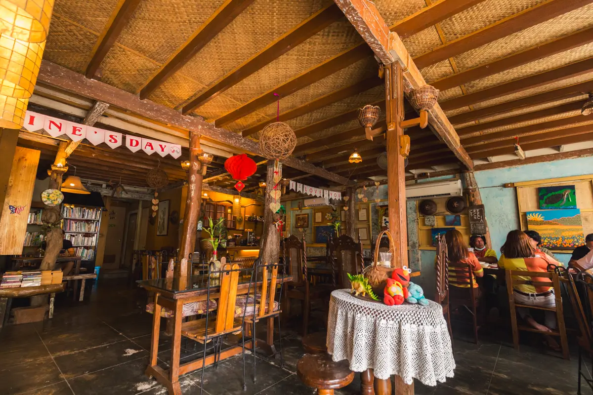 A cozy café interior with wooden beams, mismatched tables and chairs, shelves of books, wall art, and eclectic decorations inspired by Bohol. Several people sit at tables, and a crochet-covered table with ornaments is in the foreground.