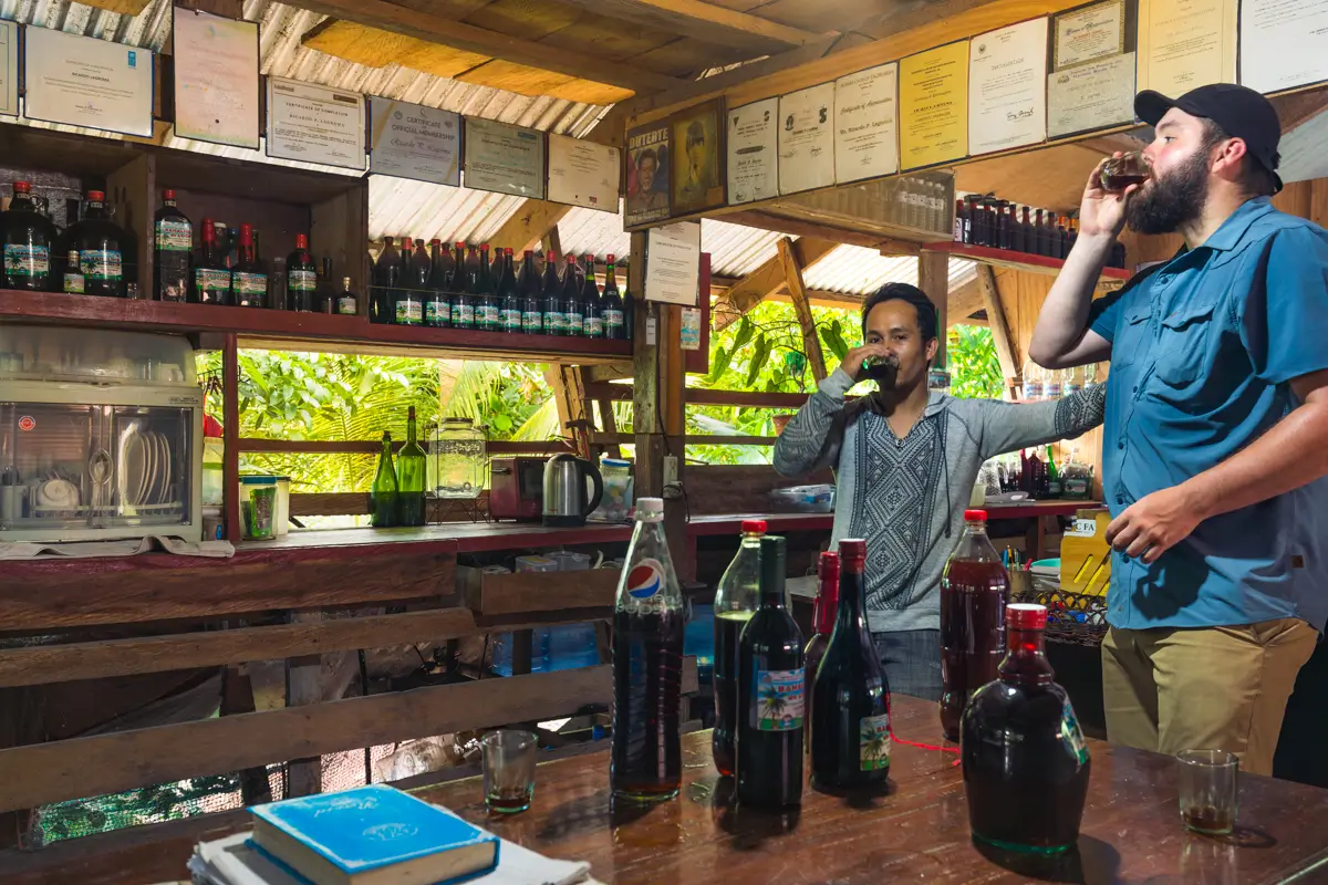 Two men stand inside a rustic Bohol bar, each drinking from a glass. Shelves behind them hold bottles and framed certificates. Multiple bottles are on the wooden counter. Greenery is visible outside through open windows.