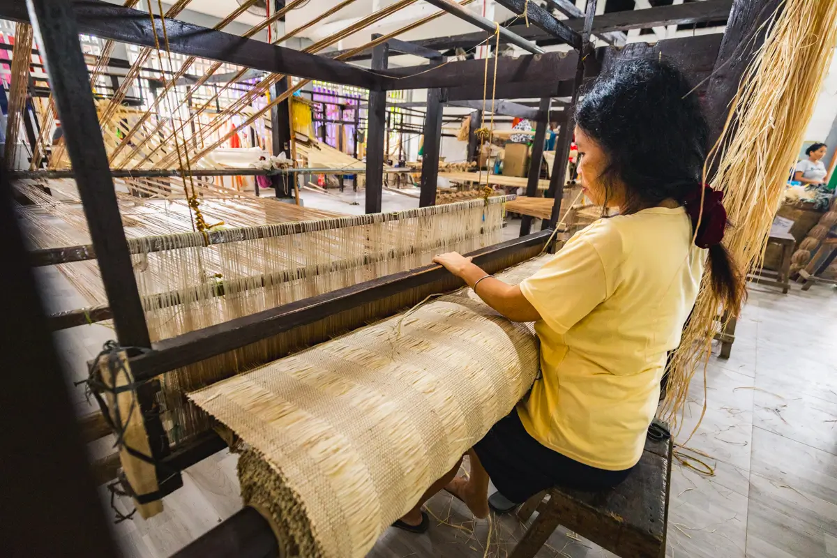 A person sits at a large wooden loom in Bohol, weaving light-colored fibers into fabric in an indoor workspace. Other people and weaving materials are visible in the background.