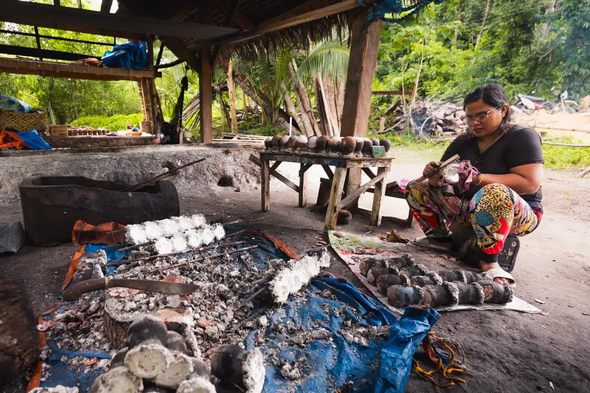 A woman in Bohol sits on the ground next to a wooden table, preparing food beside an outdoor fire pit where several charred, round items are laid out on a blue tarp and cooking tools are scattered nearby.
