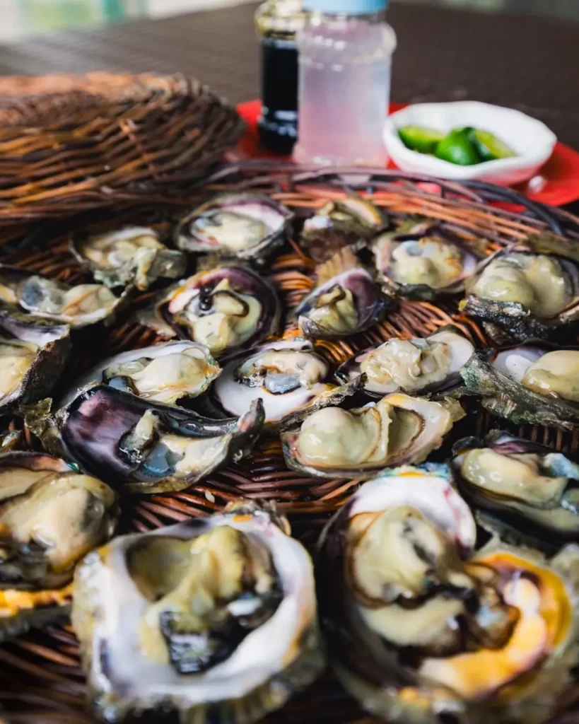A close-up of several freshly shucked oysters from Bohol arranged on a wicker tray, with a small bowl of green calamansi and bottles of condiments in the background.