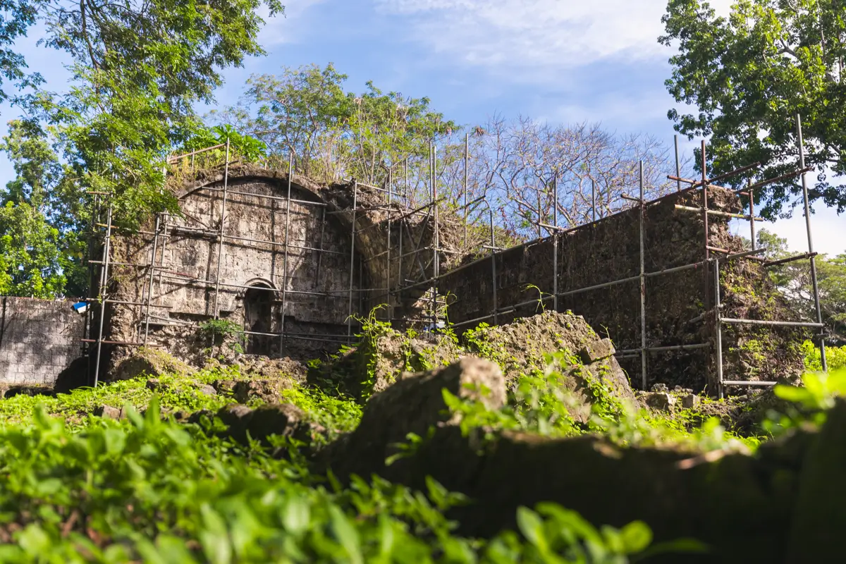 A stone building in ruins stands surrounded by green vegetation and trees in Bohol, with scaffolding set up along its walls, under a blue sky with scattered clouds.