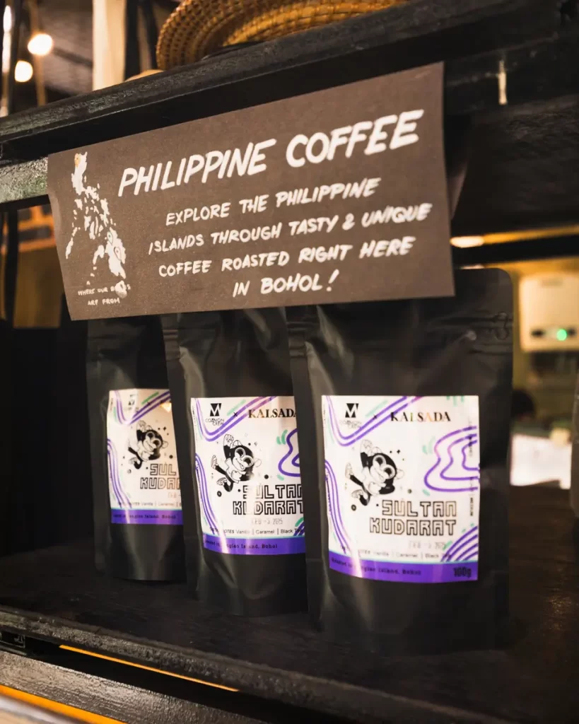 Three black bags of Philippine coffee labeled Sultan Kudarat are displayed on a shelf below a sign promoting locally roasted Bohol coffee. The bags feature a modern design with purple and white accents.