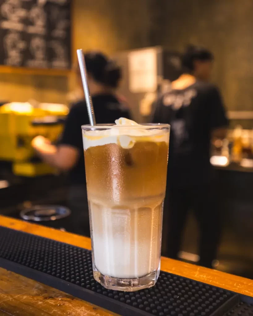 A glass of iced coffee with milk and ice cubes sits on a bar counter in a cozy Bohol coffee shop, with a metal straw. In the background, two people are blurred as they work behind the counter.