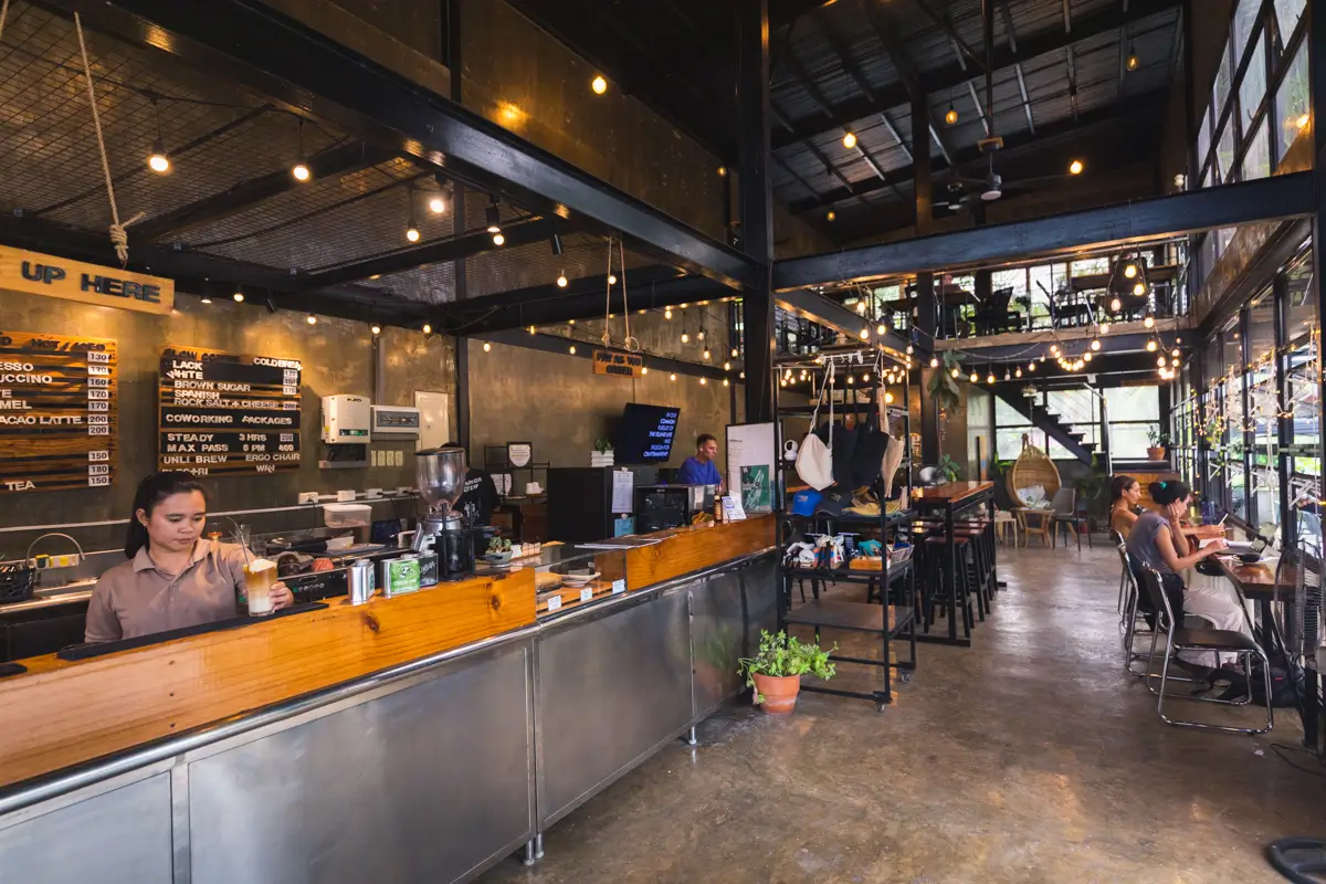 A modern coffee shop in Bohol with industrial decor features string lights, a long wooden counter, a menu board, baristas working, customers seated at tables, and a staircase leading to an upper level.