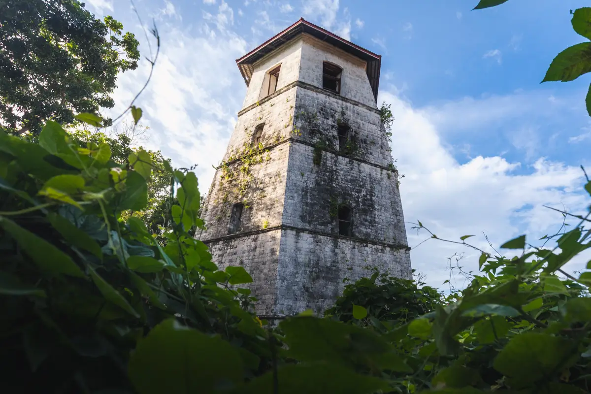 A tall, old stone tower with a red roof stands among green foliage in Bohol, viewed from a low angle against a blue sky with scattered clouds. The tower has several small windows and shows signs of age and weathering.