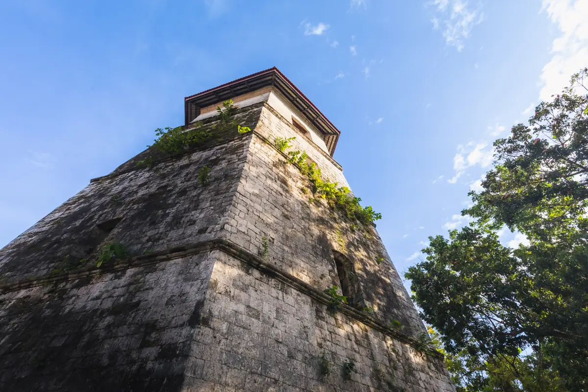 Stone tower with a square base, partially covered in small patches of greenery, photographed from a low angle against a blue Bohol sky with clouds and tree branches on the right side.