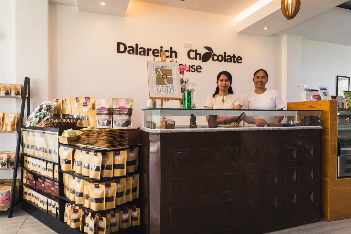 Two women stand behind the counter at Dalareich Chocolate House in Bohol, with shelves of chocolate products and bags of ingredients to the left and an award sign displayed behind the counter.