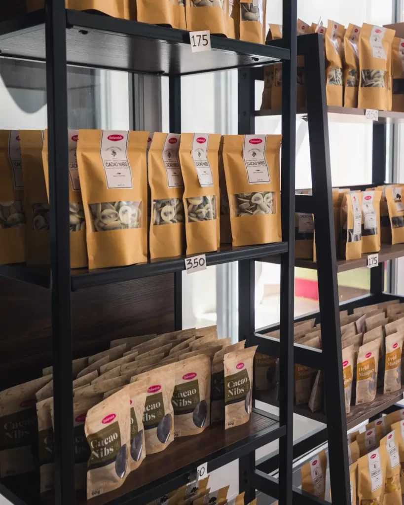 Bags of Bohol cacao nibs and other products are arranged on black metal shelves with price tags attached. A ladder leans against the shelving unit, while sunlight streams through the window in the background.