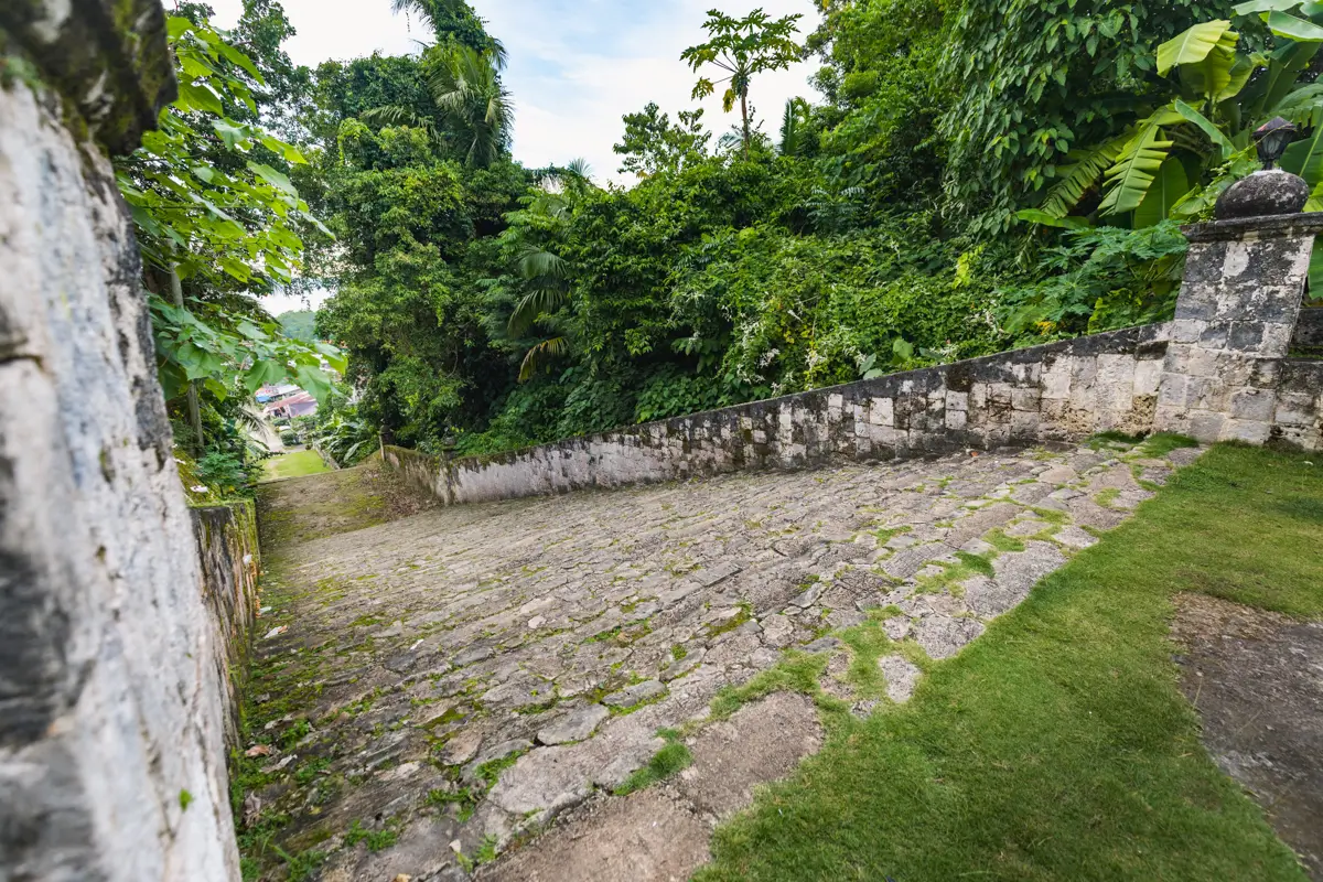 A stone-paved, sloped pathway in Bohol, bordered by old stone walls and surrounded by dense green vegetation under a partly cloudy sky.