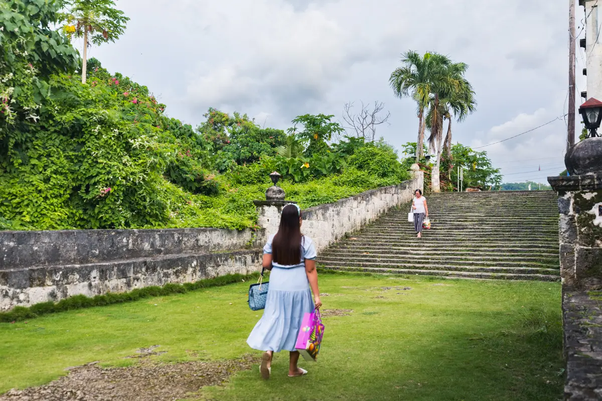 A woman in a light blue dress carrying bags walks toward stone steps surrounded by greenery and trees in Bohol. Another person stands at the top of the stairs beneath a cloudy sky.