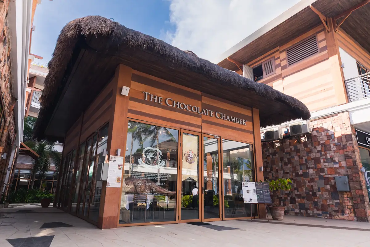 A small café called The Chocolate Chamber with glass walls and a thatched roof, nestled in Bohol’s shopping area amid stone and wood buildings. Signs and menus are displayed outside the entrance, inviting visitors to indulge.